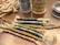 Colorful braided cords with intricate patterns are laid out on a flat surface. These cords are woven with different shades of blue, gold, and pink threads. Nearby, spools of thread in various colors, including gold and teal, are visible, as well as some thicker, natural-colored rope in the background.