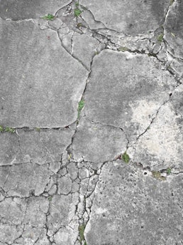 Weathered and cracked concrete surface with patches of green grass sprouting between the fissures. The texture of the concrete is rough, with various shades of gray, and some areas show signs of wear and erosion.