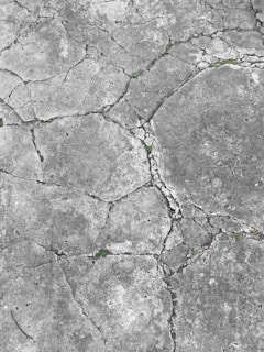 Cracked concrete surface with visible grass sprouting from the crevices, displaying various shades of gray and some patches of green.