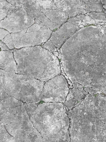 Cracked concrete surface with visible grass sprouting from the crevices, displaying various shades of gray and some patches of green.