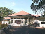 A single-story house with a red-tiled roof, cream-colored walls, and multiple windows in a serene environment. The driveway is shaded by surrounding trees, with some potted plants near the entrance.