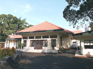 A single-story house with a red-tiled roof, cream-colored walls, and multiple windows in a serene environment. The driveway is shaded by surrounding trees, with some potted plants near the entrance.