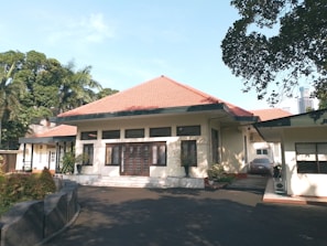 A single-story house with a red-tiled roof, cream-colored walls, and multiple windows in a serene environment. The driveway is shaded by surrounding trees, with some potted plants near the entrance.
