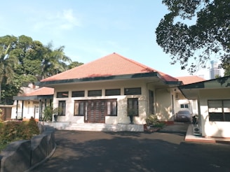 A single-story house with a red-tiled roof, cream-colored walls, and multiple windows in a serene environment. The driveway is shaded by surrounding trees, with some potted plants near the entrance.