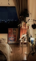 A cozy bedroom features a bed with white bedding, a wooden floor, and warm lighting. A large window reveals a night cityscape. The room is decorated with plants, artwork, and various light fixtures. A round table holds a lamp and other small items.
