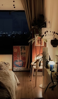 A cozy bedroom features a bed with white bedding, a wooden floor, and warm lighting. A large window reveals a night cityscape. The room is decorated with plants, artwork, and various light fixtures. A round table holds a lamp and other small items.