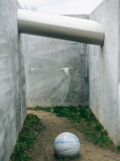 A large marble-like sphere rests on a dirt path, surrounded by concrete walls. Above, a cylindrical metal tube is embedded across the top of the walls, casting shadows on the surface.