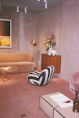 A cozy living room features a modern and stylish interior design. It includes a beige velvet sofa, a glass coffee table, and a uniquely designed black and white striped chair. A wooden cabinet with a brass lamp and a floral arrangement adds a touch of elegance. The room has a neutral color palette with soft lighting.
