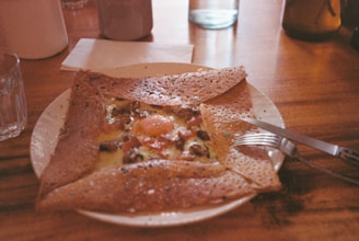 A crepe with an egg and toppings such as mushrooms and herbs on a white plate, placed on a wooden table alongside a fork and knife. The ambiance is warm with diffused lighting, and there are cups and glass bottles surrounding the dish.