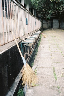 A row of ceramic sinks lined up against a metal railing, with two straw brooms leaning against the wall in an outdoor setting. The ground is paved with square tiles, and there is greenery in the background.