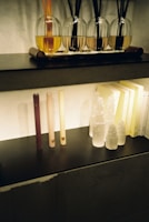 A shelf features a row of reed diffusers with glass bottles containing colored liquids and black reeds extending out of them. Below, there are several tall, thin candles of varying colors, alongside decorative crystal-like objects resembling towers. Soft lighting illuminates the setup, casting gentle shadows behind the objects.