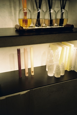 A shelf features a row of reed diffusers with glass bottles containing colored liquids and black reeds extending out of them. Below, there are several tall, thin candles of varying colors, alongside decorative crystal-like objects resembling towers. Soft lighting illuminates the setup, casting gentle shadows behind the objects.