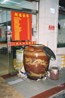 A large decorative jar with a dragon design is placed outside a restaurant entrance. The jar is accompanied by a smaller container with a brush. A red menu with yellow text displaying a list of food items and prices is visible on the glass door. The interior of the restaurant is partially visible.
