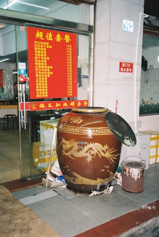 A large decorative jar with a dragon design is placed outside a restaurant entrance. The jar is accompanied by a smaller container with a brush. A red menu with yellow text displaying a list of food items and prices is visible on the glass door. The interior of the restaurant is partially visible.