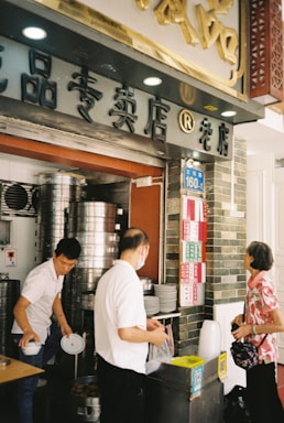 A busy street food kitchen with steaming equipment and three people interacting. The shop's facade includes Chinese characters and a numbered street sign. One person is handling stacks of plates, while the other appears to be serving food. An elderly woman is waiting, carrying a handbag.