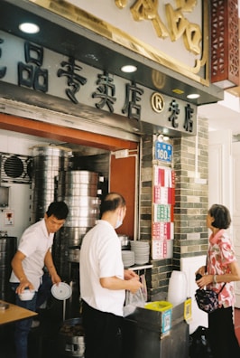 A busy street food kitchen with steaming equipment and three people interacting. The shop's facade includes Chinese characters and a numbered street sign. One person is handling stacks of plates, while the other appears to be serving food. An elderly woman is waiting, carrying a handbag.