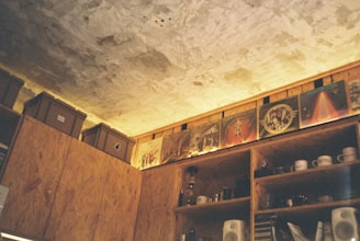 A rustic and cozy interior with wooden shelves and cabinets. Several record albums are displayed along a beam near the ceiling. Some boxes are placed on top of the cabinets, and a variety of cups and kitchen items are stored in the open shelves.
