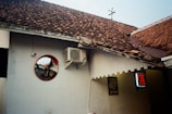 The image features a section of a building with white walls and a brown tiled roof. An air conditioning unit is mounted high on the wall. A circular mirror with a red frame reflects part of the building and a street view. There's a partially visible illuminated sign with red lettering to the right, and part of a framed poster is also visible on the wall.