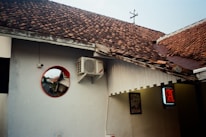 The image features a section of a building with white walls and a brown tiled roof. An air conditioning unit is mounted high on the wall. A circular mirror with a red frame reflects part of the building and a street view. There's a partially visible illuminated sign with red lettering to the right, and part of a framed poster is also visible on the wall.