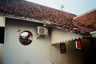 The image features a section of a building with white walls and a brown tiled roof. An air conditioning unit is mounted high on the wall. A circular mirror with a red frame reflects part of the building and a street view. There's a partially visible illuminated sign with red lettering to the right, and part of a framed poster is also visible on the wall.