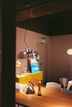 A modern interior setting with a focus on a wooden table adorned with a stainless steel kettle and an orange flashlight. A yellow cabinet holds colorful objects and artwork. A contemporary lamp hangs above, casting light on the arrangement. The room features soft lighting, creating a cozy atmosphere, with a visible exit sign on a door in the background.