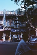 A group of people are gathered outside a row of small shops on a tree-lined street. The building features air conditioning units on the facade and has a rustic, old-world architecture. Shadows from trees create patterns on the ground and building exterior, indicating a sunny day.