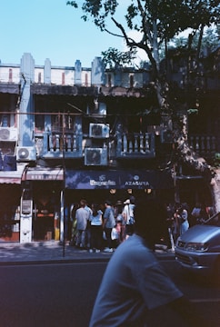 A group of people are gathered outside a row of small shops on a tree-lined street. The building features air conditioning units on the facade and has a rustic, old-world architecture. Shadows from trees create patterns on the ground and building exterior, indicating a sunny day.