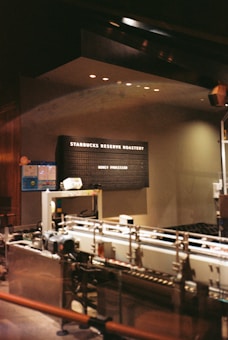A dimly lit industrial-style interior featuring machinery and equipment. A black signboard displays the text 'Starbucks Reserve Roastery' and 'Honey Processed'. The setting suggests a specialized coffee processing area, with metallic pipes and a modern, sophisticated ambiance.