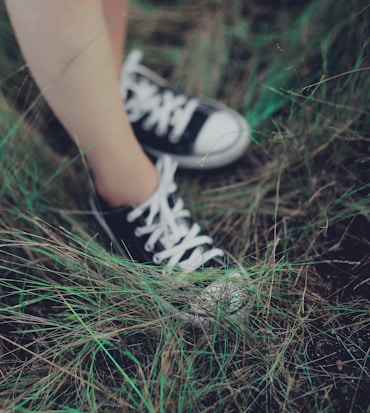 A pair of black and white sneakers on a person's feet standing amidst dense green grass. The grass appears wild and slightly untamed, giving a sense of nature and casual outdoor exploration.
