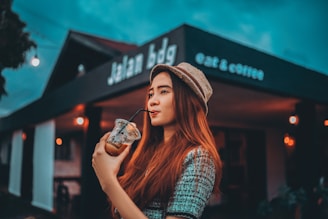 A person with long hair and a hat is sipping a drink from a plastic cup with a straw, standing in front of a cafe or restaurant. The establishment has a sign that reads 'Jalan bdg' and 'eat & coffee'. The background is slightly blurred with visible warm-toned lights.