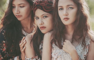 Three women are closely grouped together, displaying a somber and contemplative expression. They have long wavy hair, and one woman wears a patterned headscarf. The clothing is intricate with lace and floral patterns.