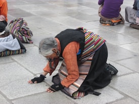 An individual dressed in traditional clothing is kneeling on a stone pavement. The person's attire includes a colorful striped apron and an orange shirt. The head is bowed, and hands are placed on the ground, suggesting prayer or meditation. Others in similar attire are seen in the background.