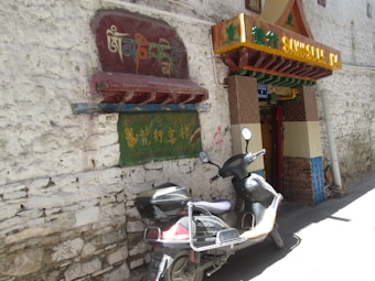 A rustic wall with textured stones hosts colorful Tibetan and other Asian scripts painted on a sign. Below this, a black scooter is parked, partially shadowed, at the entrance to a small structure with a distinctive, decorative awning. The entrance is adorned with traditional patterns and bright colors, providing a vibrant contrast to the muted tones of the wall.