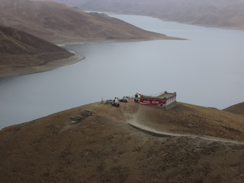 A remote building with a large red advertising sign is located on a barren hill overlooking a vast body of water. Several people and a few vehicles are gathered near the building. The landscape is characterized by rugged, rolling hills and the sky is overcast, giving the scene a desolate and isolated feel.
