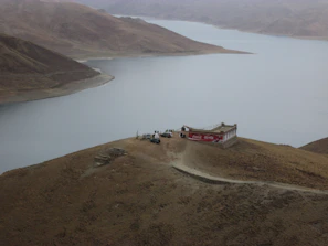 A remote building with a large red advertising sign is located on a barren hill overlooking a vast body of water. Several people and a few vehicles are gathered near the building. The landscape is characterized by rugged, rolling hills and the sky is overcast, giving the scene a desolate and isolated feel.