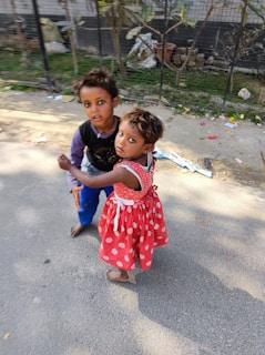 Two children stand on a sidewalk. One child is wearing a red dress with white polka dots, and the other is dressed in a black top with a purple shirt underneath and blue pants. They are barefoot and appear to be holding hands. The background shows a fence and some foliage, with scattered items on the ground.