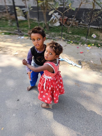 Two children stand on a sidewalk. One child is wearing a red dress with white polka dots, and the other is dressed in a black top with a purple shirt underneath and blue pants. They are barefoot and appear to be holding hands. The background shows a fence and some foliage, with scattered items on the ground.