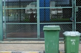 Two green garbage bins stand on a paved sidewalk in front of a chain-link fence. The background shows a patio area with glass windows reflecting buildings and greenery.