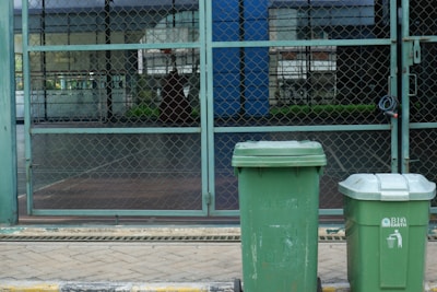 Two green garbage bins stand on a paved sidewalk in front of a chain-link fence. The background shows a patio area with glass windows reflecting buildings and greenery.