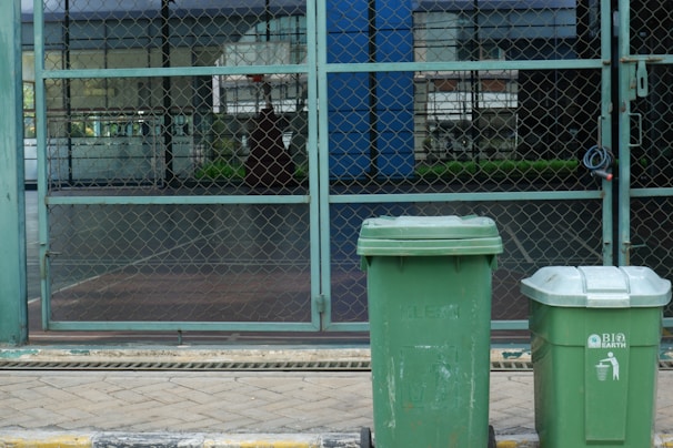 Two green garbage bins stand on a paved sidewalk in front of a chain-link fence. The background shows a patio area with glass windows reflecting buildings and greenery.