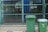 Two green garbage bins stand on a paved sidewalk in front of a chain-link fence. The background shows a patio area with glass windows reflecting buildings and greenery.