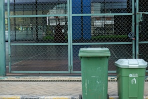 Two green garbage bins stand on a paved sidewalk in front of a chain-link fence. The background shows a patio area with glass windows reflecting buildings and greenery.
