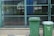 Two green garbage bins stand on a paved sidewalk in front of a chain-link fence. The background shows a patio area with glass windows reflecting buildings and greenery.