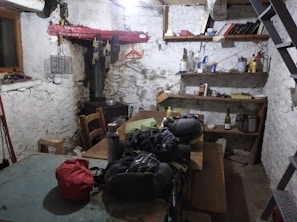 A rustic and cluttered room with raw, plastered walls and simple wooden furniture including a large communal table. There are various objects on the shelves, like books, bottles, and household items. The space gives an impression of an old storage or utility room, with a visible staircase and basic amenities.