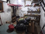 A rustic and cluttered room with raw, plastered walls and simple wooden furniture including a large communal table. There are various objects on the shelves, like books, bottles, and household items. The space gives an impression of an old storage or utility room, with a visible staircase and basic amenities.