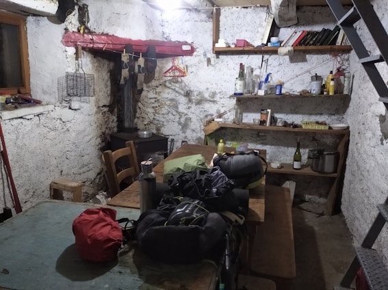 A rustic and cluttered room with raw, plastered walls and simple wooden furniture including a large communal table. There are various objects on the shelves, like books, bottles, and household items. The space gives an impression of an old storage or utility room, with a visible staircase and basic amenities.
