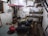 A rustic and cluttered room with raw, plastered walls and simple wooden furniture including a large communal table. There are various objects on the shelves, like books, bottles, and household items. The space gives an impression of an old storage or utility room, with a visible staircase and basic amenities.