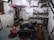 A rustic and cluttered room with raw, plastered walls and simple wooden furniture including a large communal table. There are various objects on the shelves, like books, bottles, and household items. The space gives an impression of an old storage or utility room, with a visible staircase and basic amenities.