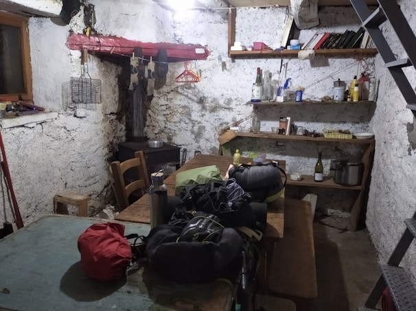 A rustic and cluttered room with raw, plastered walls and simple wooden furniture including a large communal table. There are various objects on the shelves, like books, bottles, and household items. The space gives an impression of an old storage or utility room, with a visible staircase and basic amenities.