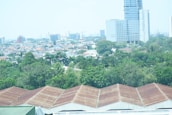 An urban landscape with a blend of natural and architectural elements. In the foreground, several large industrial buildings with brownish roofs are visible, surrounded by lush green trees and vegetation. Behind the greenery, a densely packed residential area with various houses can be seen, leading up to taller modern buildings and skyscrapers in the background.